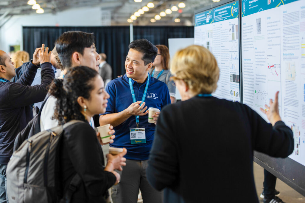 A person in a polo shirt speaks to three other people in front of a poster at a conference