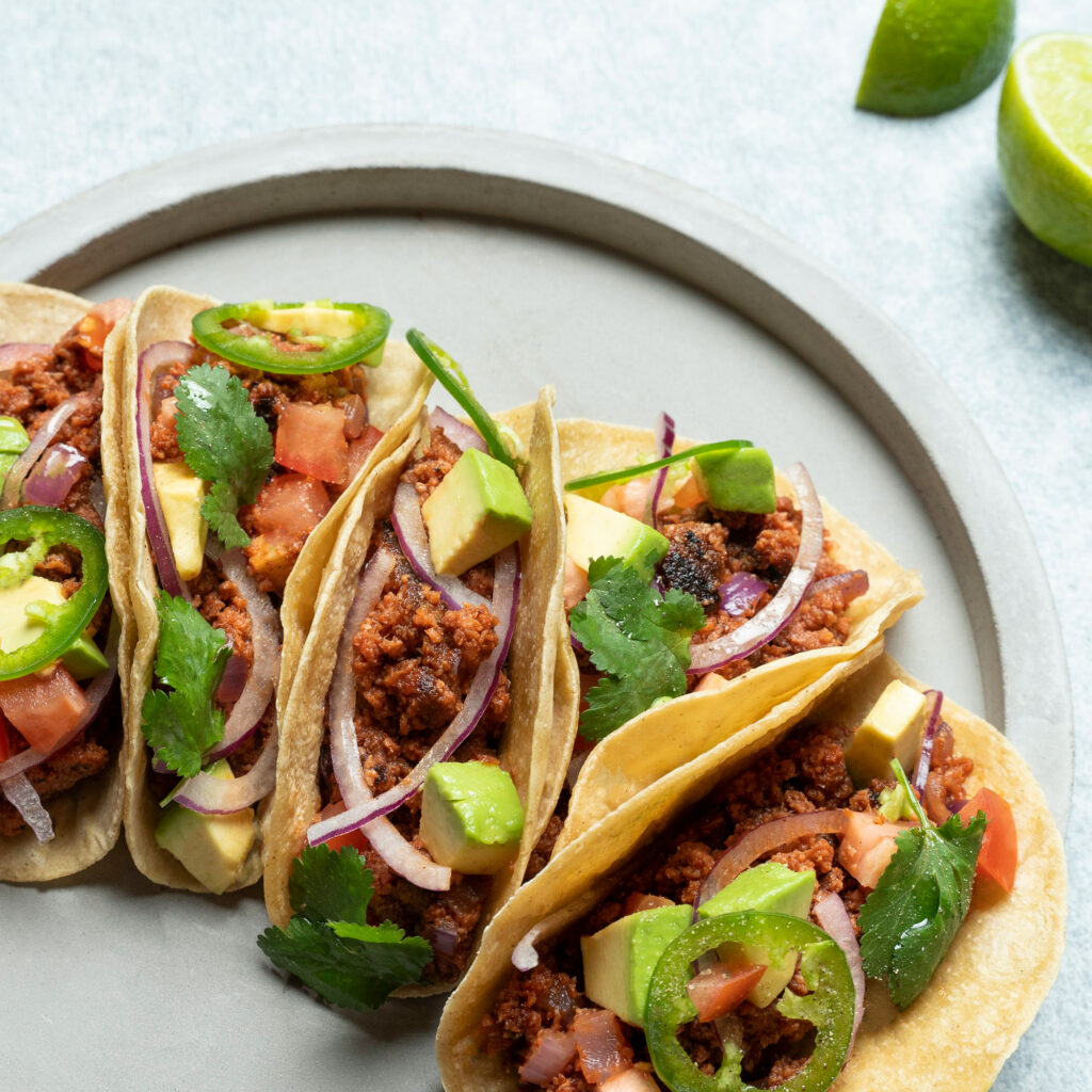 Five tacos on a gray plate with garnishes and lime wedges. Photo credit: magic valley.